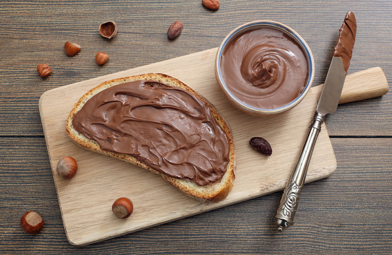 A cutting board with a knife, slice of bread with Choco spread on top beside a bowl filled with Choco spread.