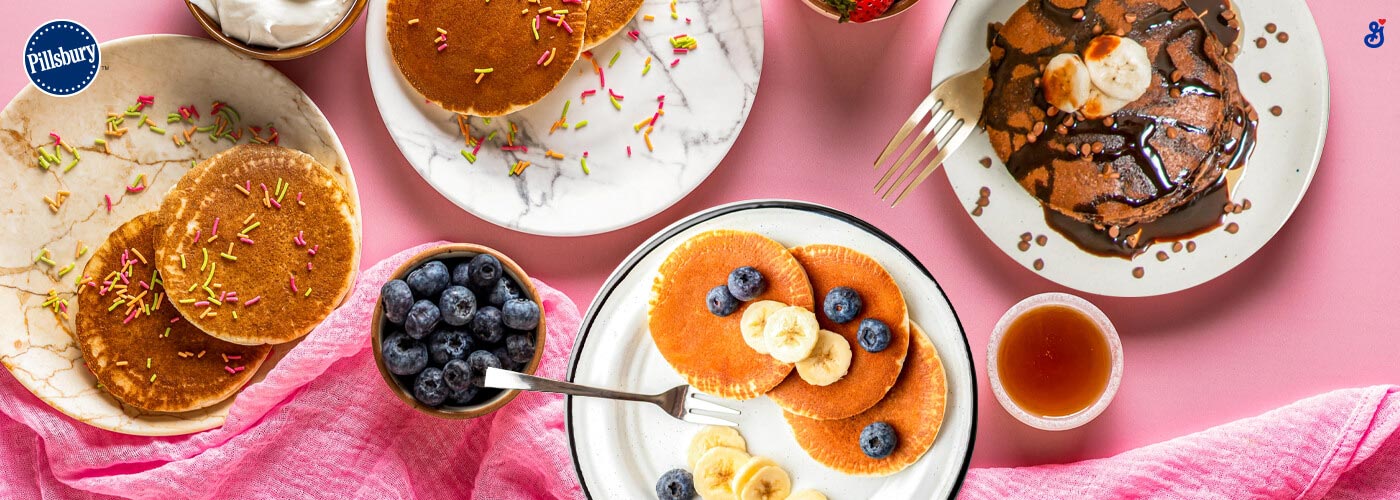 A mix of white plates on a pink table. Pancakes, blueberries and syrup with gold silverware