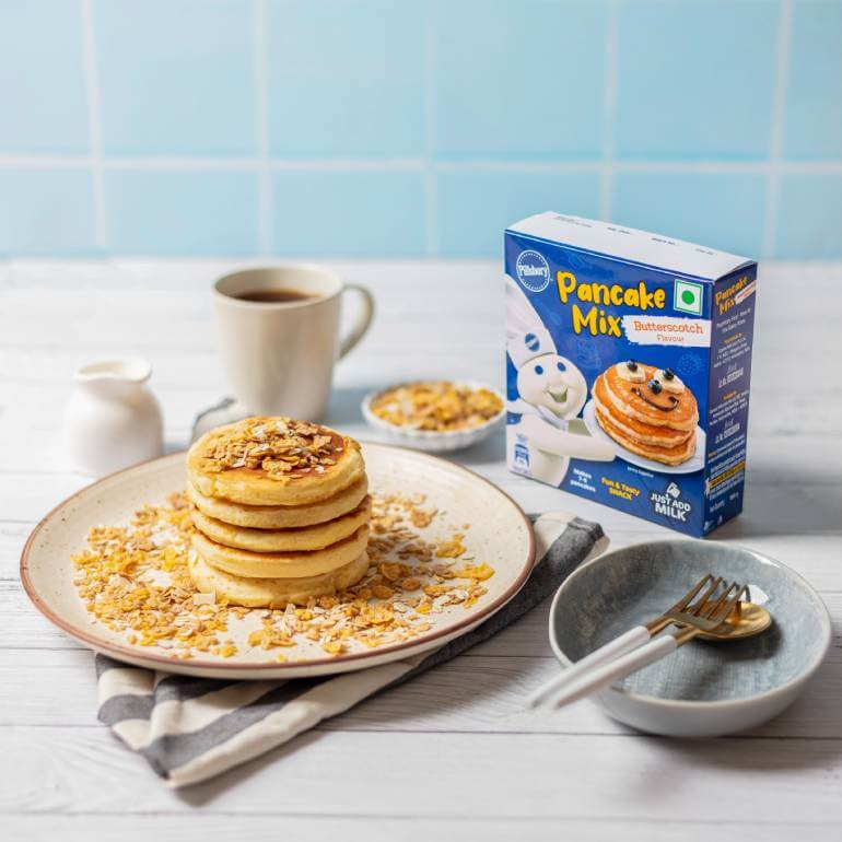 A plate of Butterscotch pancakes with a cup of coffee, silverware and a napkin. Also includes a box of Butterscotch Pancakes Mix.